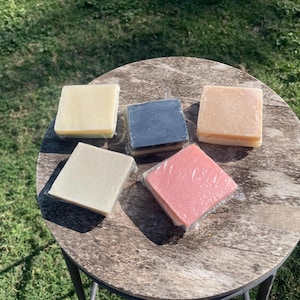 May include: Five square bars of handmade soap in various colors, including cream, blue, peach, and pink, are arranged on a round, weathered wooden table. The soaps are individually wrapped in clear plastic, and the table sits on a metal frame, with green grass in the background.