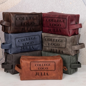 May include: A stack of leather toiletry bags in various colors, including brown, burgundy, and blue. Each bag has a rectangular label with the text "COLLEGE LOGO JULIA". The bags have a zipper closure and a side handle.