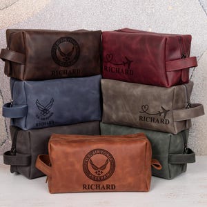 May include: A collection of rectangular leather toiletry bags in various colors including brown, burgundy, blue, gray, and green. Each bag features a side handle, a zipper closure, and custom engravings of the U.S. Air Force logo or an airplane with a heart.