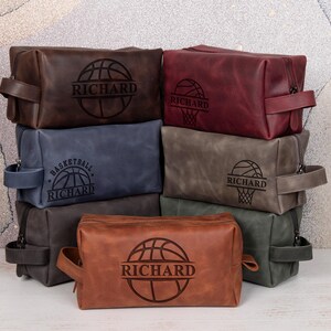May include: A collection of personalized leather toiletry bags in various colors, including brown, burgundy, and blue. Each bag features a basketball design and the name "Richard" engraved on the front. The bags have a rectangular shape with a zipper closure and a handle.