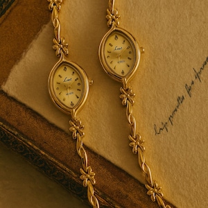 May include: Two gold-coloured wristwatches with oval faces and floral-linked bands. The watch faces are cream-coloured with black numerals and hands, and the word "Loot" is printed on the face. The watches rest on a textured, tan surface.