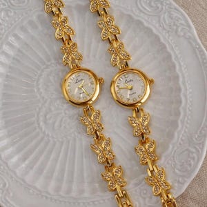 Puede incluir: Dos relojes de pulsera dorados con eslabones en forma de mariposa. Las esferas son redondas con fondo blanco y agujas doradas. Las correas están adornadas con diseños de mariposas y los relojes se exhiben en un plato blanco.