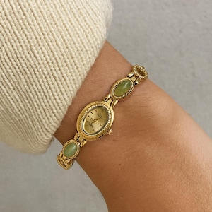 May include: Gold wristwatch with an oval face and a matching bracelet. The watch has a light-coloured face with gold hands and markers. The bracelet features oval-shaped green stones. The watch is worn on a wrist, with a cream-coloured sweater sleeve in the background.