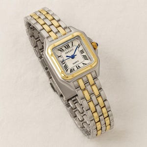 Puede incluir: Un reloj de pulsera plateado y dorado con esfera cuadrada. El reloj presenta números romanos, agujas azules y la palabra "GREALY" en la esfera. La correa es una combinación de eslabones plateados y dorados.