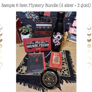 Op de afbeelding: Een verzameling mystieke items, waaronder een zwarte waterfles met een bloemen slangenontwerp, een rode oogmaskerdoos met de tekst "I'm Not A Morning Person" en een tarotkaartspel. Een zwart Ouija-bord en een ketting zijn ook inbegrepen.