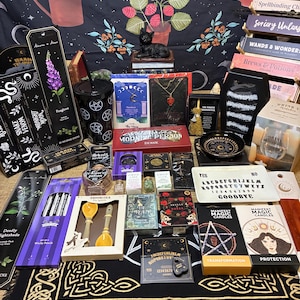 Puede incluir: Una colección de artículos místicos y de brujería, que incluyen incienso, velas, libros y joyas. Los artículos están dispuestos sobre una tela negra con diseños de nudos celtas dorados. Una estatua de gato negro se encuentra sobre libros.