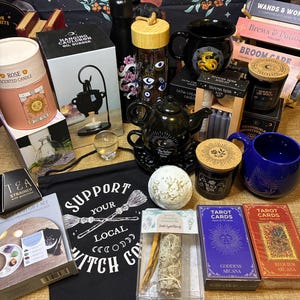 Op de afbeelding: Een verzameling hekserij- en spirituele items, waaronder kaarsen, tarotkaarten, een theepot, mokken en een banner met de tekst "Support Your Local Witch Co."