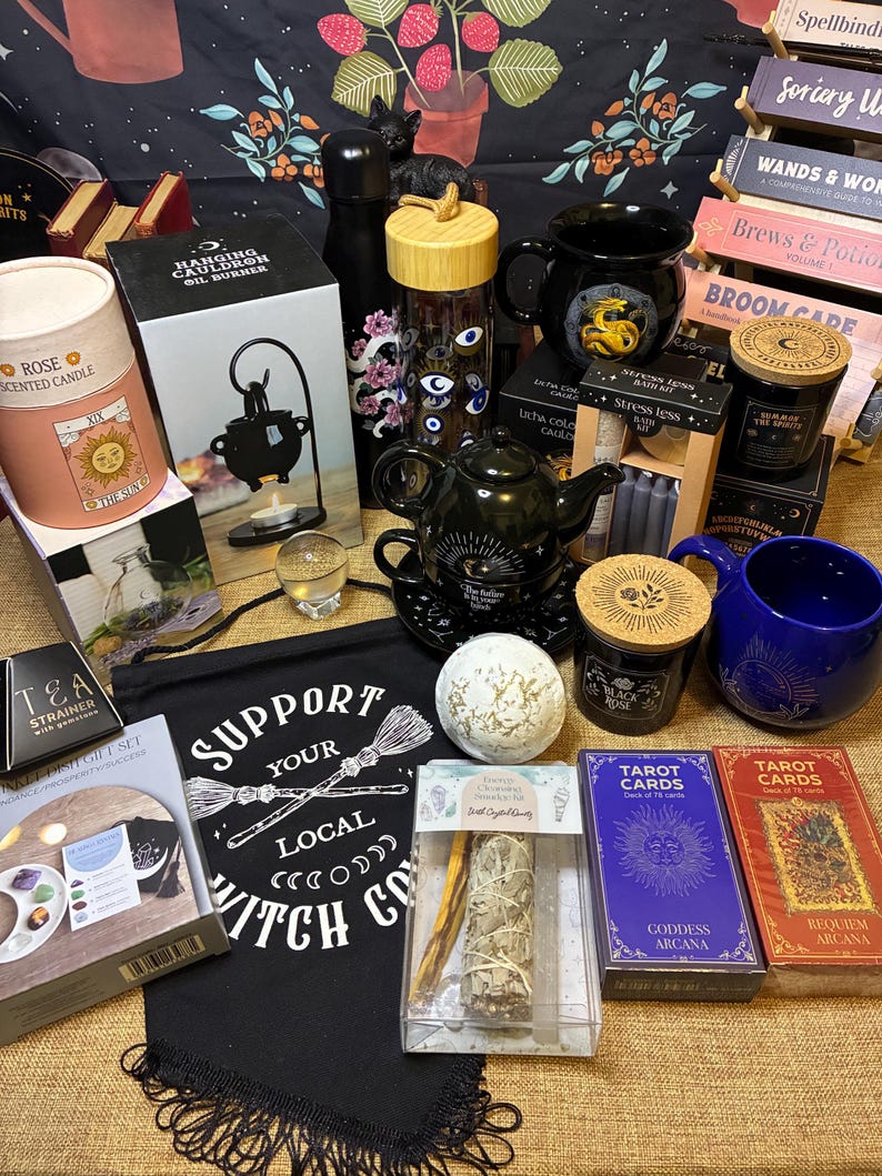 Op de afbeelding: Een verzameling hekserij- en spirituele items, waaronder kaarsen, tarotkaarten, een theepot, mokken en een banner met de tekst "Support Your Local Witch Co."