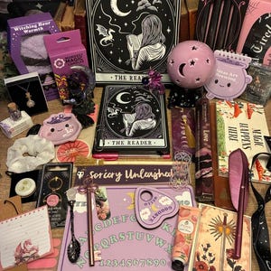 Puede incluir: Una colección de papelería y accesorios que incluye un diario negro titulado The Reader, un tablero ouija morado, una lámpara de luna rosa, una crema de manos de melocotón, un bolígrafo de pluma, un bolígrafo de llave, una cuchara negra y tarjetas.