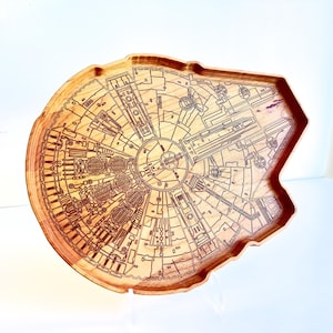 May include: A wooden tray shaped like the Millennium Falcon starship, featuring intricate laser-engraved mechanical details on its surface. The natural light brown wood grain is visible throughout the tray, which has a raised edge and a flat base.