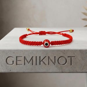 May include: A red braided cord bracelet featuring a central glass evil eye bead with a white, blue, and black iris. The bracelet has an adjustable sliding knot closure with small gold-toned metal beads at the ends of the cord strings.
