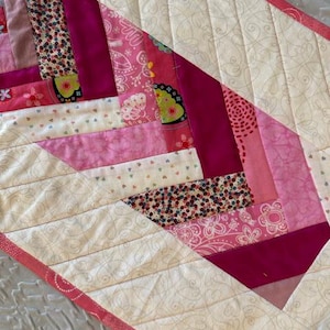 May include: A handmade quilt featuring a patchwork design in shades of pink, fuchsia, and cream. The quilt has a geometric pattern with various floral and patterned fabrics. The edges are finished with a pink border and detailed stitching.