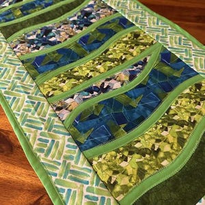 May include: A quilted table runner with a vibrant design. The runner features wavy bands of green, blue, and patterned fabrics, bordered by a green and white geometric print. The stitching is visible, adding texture to the piece. The runner is designed to be placed on a table.