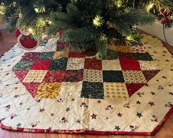 May include: A quilted Christmas tree skirt with a patchwork design. The center features red, green, and gold squares. The outer border is cream-colored with red stars and bird motifs. The skirt is under a decorated Christmas tree.