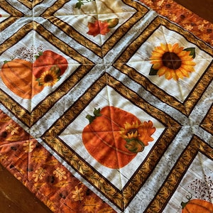 May include: A quilted table runner with a fall theme. It features a white background with brown, orange, and yellow pumpkins, sunflowers, and leaves. The runner has a brown and orange border with a leaf pattern.