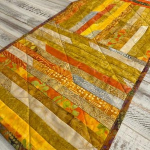 May include: A rectangular quilted table runner featuring a diagonal patchwork design in shades of mustard yellow, burnt orange, cream, and olive green. The fabric pieces display various textures and patterns, finished with diamond-shaped stitching.