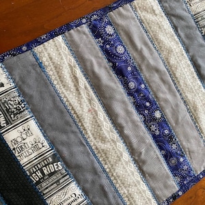 May include: A quilted table runner with a patchwork design. The runner features vertical stripes in shades of gray, blue, and white. Some stripes have text, and others have snowflake patterns. The edges are finished with blue stitching.