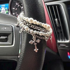 May include: A layered bracelet set with faux pearls and silver-tone chains and charms, including crosses and crescent moons. The bracelets are draped over a black steering wheel.