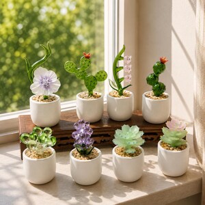 Op de afbeelding: Een verzameling glazen sculpturen van vetplanten en bloemen in kleine witte potten. De sculpturen omvatten verschillende vormen en kleuren, zoals groene cactussen, paarse bloemen en roze bloesems. De potten zijn gevuld met kleine kiezels.