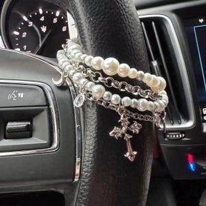May include: A layered bracelet set with faux pearls and silver-tone chains and charms. The charms include a cross, a crescent moon, and a small pendant. The bracelets are draped over a black steering wheel.