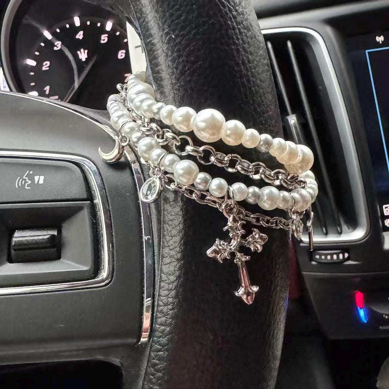 May include: A layered bracelet set with faux pearls and silver-tone chains and charms. The charms include a cross, a crescent moon, and a small pendant. The bracelets are draped over a black steering wheel.