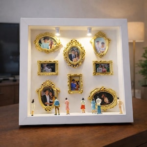 Może przedstawiać: Białe pudełko z dziewięcioma złotymi ramkami o różnych rozmiarach, zawierającymi zdjęcia. Małe figurki stoją przed zdjęciami. Pudełko jest oświetlone przez dwa małe światła.