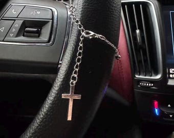 May include: A silver-colored cross pendant hanging from a chain, attached to a car's steering wheel. The cross is a simple, classic design. The steering wheel is black leather.