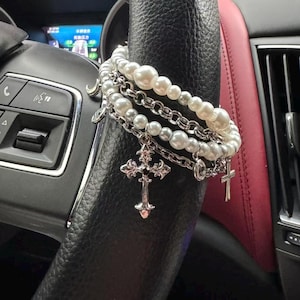 May include: A layered bracelet featuring faux pearls and silver-toned chains. The bracelet includes multiple strands of varying pearl sizes and chain links, with dangling cross charms. The jewelry is displayed on a black steering wheel.