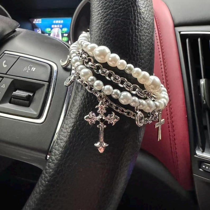 May include: A layered bracelet featuring faux pearls and silver-toned chains. The bracelet includes multiple strands of varying pearl sizes and chain links, with dangling cross charms. The jewelry is displayed on a black steering wheel.
