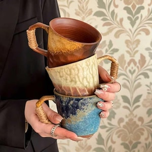 May include: A stack of three ceramic mugs with textured surfaces and rope-wrapped handles. The top mug is a deep brown, the middle mug is a creamy beige with vertical ridges, and the bottom mug features a mottled blue and brown glaze finish.