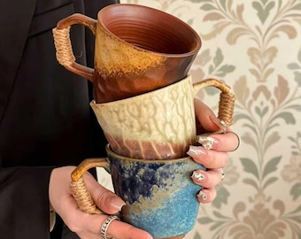 May include: A stack of three ceramic mugs with textured surfaces and rope-wrapped handles. The top mug is a deep brown, the middle mug is a creamy beige with vertical ridges, and the bottom mug features a mottled blue and brown glaze finish.