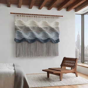 May include: A large, woven wall hanging with a wave-like design in shades of white, gray, and blue. The textile is suspended from a wooden rod and has long, gray fringe. A wooden chair and a rug are in the foreground.