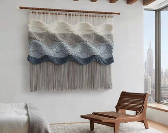 May include: A large, woven wall hanging with a wave-like design in shades of white, gray, and blue. The textile is suspended from a wooden rod and has long, gray fringe. A wooden chair and a rug are in the foreground.