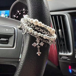 May include: A collection of pearl and chain bracelets with silver cross charms draped over a black steering wheel. The bracelets feature varying sizes of white pearls and delicate silver chains. The crosses are ornate and detailed.
