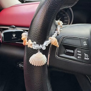 Könnte beinhalten: Ein dekorativer Autoanhänger mit einem Muschelanhänger und einer Kette aus Muscheln, klaren Perlen und korallenfarbenen Steinen. Der Anhänger ist an einem schwarzen Lenkrad befestigt.