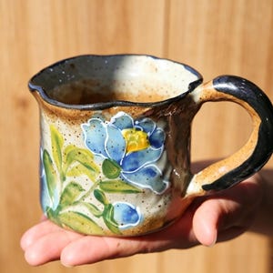 Puede incluir: Una taza de cerámica con acabado beige moteado, decorada con una flor azul pintada a mano con centro amarillo y hojas verdes. La taza presenta un borde irregular y ondulado, además de un asa gruesa de color marrón oscuro con acabado brillante.