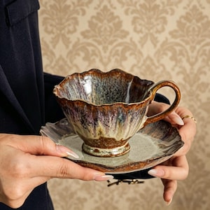 Puede incluir: Un juego de taza y platillo de cerámica con un diseño de borde único y ondulado. La taza y el platillo presentan una mezcla de tonos marrones, morados y verdes, creando un efecto veteado. El asa es curvada y el platillo tiene un borde festoneado a juego.