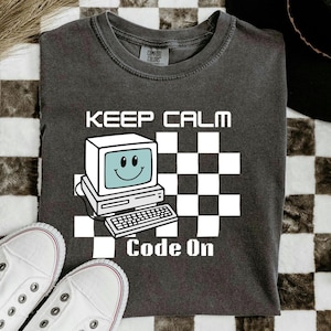 May include: Dark gray t-shirt with the words "KEEP CALM Code On" and a retro computer graphic. The computer has a smiling face on the screen. White sneakers are at the bottom of the image.