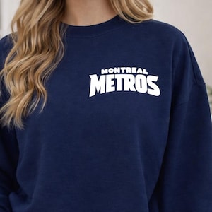 May include: Navy blue sweatshirt with the white text "MONTREAL METROS" across the chest. The sweatshirt is a crew neck style and is made of a soft material. The person in the image is wearing blue jeans.