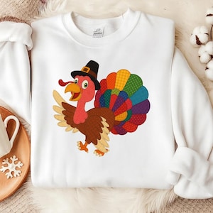 Könnte beinhalten: Weißes Sweatshirt mit einer Cartoon-Truthahn, der einen Pilgerhut trägt. Der Truthahn hat eine rote Wamme, einen gelben Schnabel und einen bunten Schwanz. Das Design ist auf der Vorderseite des Sweatshirts zentriert und macht es zu einem festlichen und auffälligen Stück.