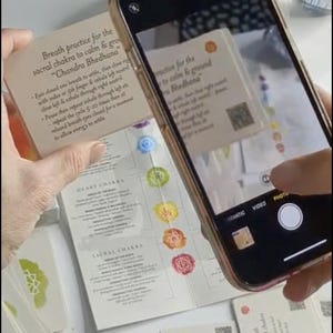 May include: A smartphone, in a gold case, is used to photograph a stack of cards. The cards contain text about yoga practices and chakra information. The cards have colourful chakra symbols and QR codes. The text includes 'Chandra Bhedhana'.