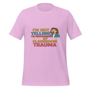 May include: A light pink t-shirt with the text "I'M NOT YELLING I'M PROCESSING MY CLASSROOM TRAUMA" in a colorful design. The graphic includes an illustration of a woman with brown hair and a blue blazer.