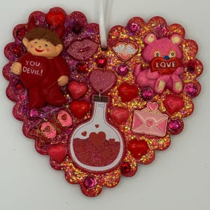 May include: Heart-shaped ornament with a glittery red and pink design. Features a red figure with the text "YOU DEVIL!", a pink teddy bear holding a heart, and a potion bottle. Embellished with hearts and gems.