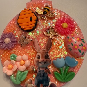 May include: A round, glittery ornament with a pink background. It features a cartoon rabbit, flowers, a beehive, a bee, butterflies, and an Easter egg. A white ribbon is attached for hanging.