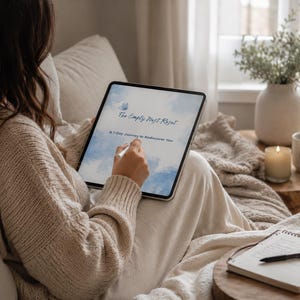 Könnte beinhalten: Eine Person sitzt auf einem Sofa und hält ein Tablet mit dem Titel "The Empty Nest Reset" und dem Untertitel "A 7-Day Journey to Rediscover You". Ein Notizbuch und ein Stift liegen auf einem Holztisch, zusammen mit einer Kerze und einer Tasse mit dem Wort "peace".