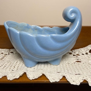 May include: A light blue ceramic planter with a unique, curved design. The planter has a scalloped edge and rests on three small feet. A decorative swirl detail is on one side. The planter is displayed on a wooden surface.