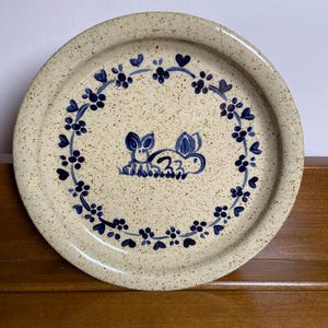 May include: A round ceramic plate with a speckled beige finish. The center features a hand-painted blue line drawing of a rabbit sitting in grass. A circular border of blue painted flowers and small heart shapes frames the central rabbit design.