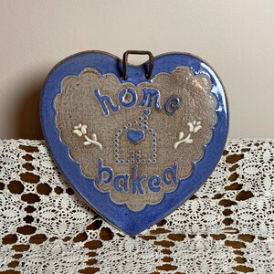 May include: Heart-shaped ceramic wall hanging with a blue border and a brown center. The words "home baked" are written in blue script. The design includes a house and floral accents. A metal loop is attached for hanging.