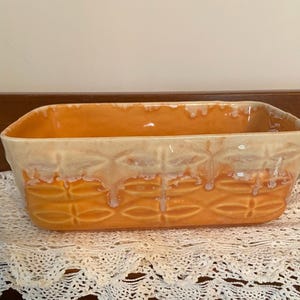 May include: An orange ceramic rectangular baking dish with a textured pattern. The top edge has a cream-colored glaze that drips down the sides. The dish is sitting on a white lace tablecloth.