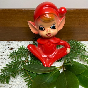 May include: A glossy red ceramic elf figurine featuring a pointed hat with a pom-pom, large pointed ears, blue eyes, and a small mouth. The elf is depicted in a seated position with legs crossed, wearing a red collar and a matching red outfit.